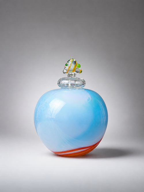A luminous handmade art glass vessel in soft light blue with orange and white accents. This one-of-a-kind lidded vessel by Jon Oaks is available from Raku Gallery in Jerome, Arizona. Perfect for collectors, shelf styling, and unique home decor.
