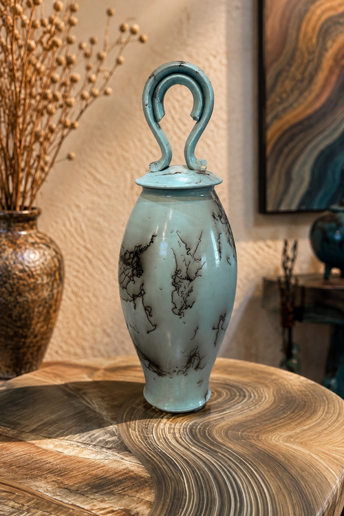 Handmade soft blue horsehair pottery vessel with lid displayed on a wood table