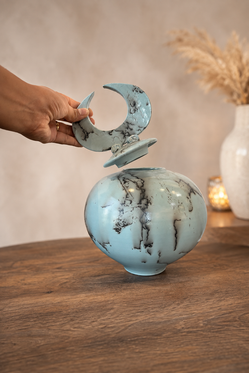 Hand lifting the crescent lid from a light blue horsehair pottery vessel