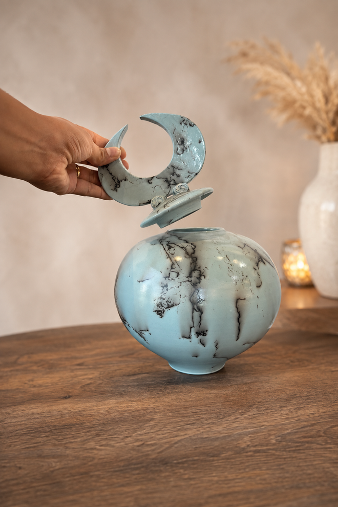 Hand lifting the crescent lid from a light blue horsehair pottery vessel